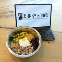 Futtrenapf mit Fleisch, Gemüse, Kartoffeln und Zusätzen vor einem Laptop mit Barky Bones-Logo